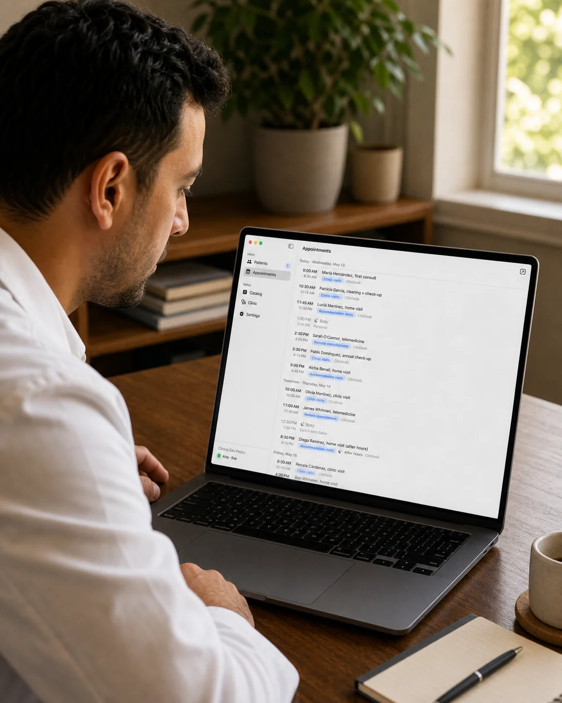 El Dr. Zavala en el escritorio de su consultorio, inclinado para leer la pantalla de su MacBook, luz suave de la tarde