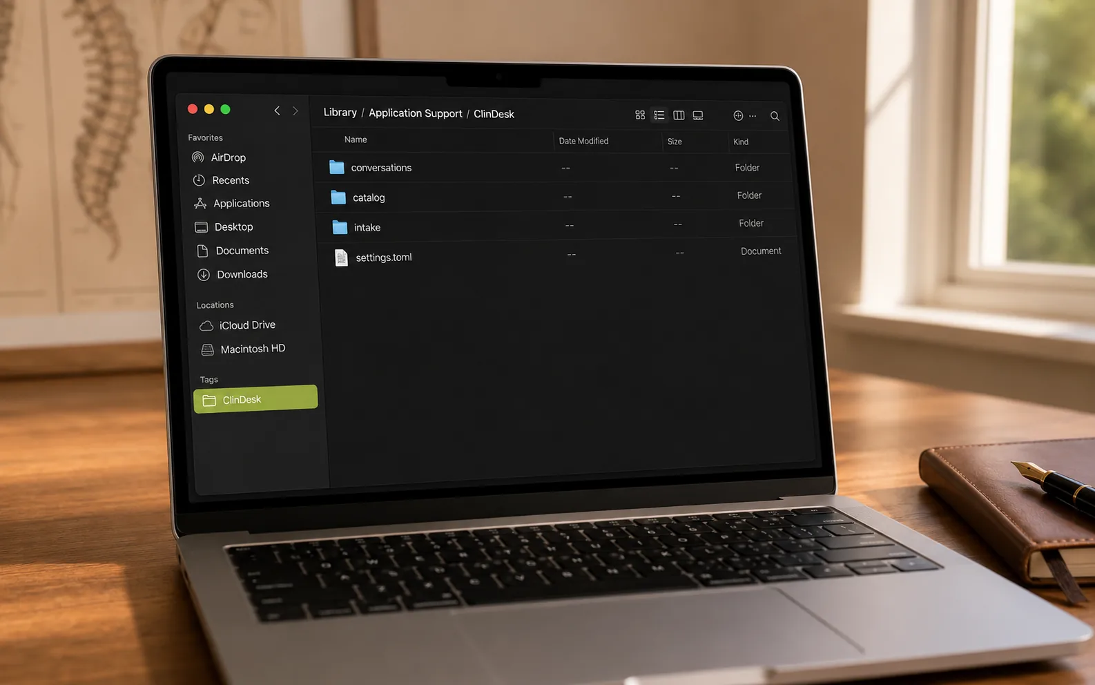 MacBook on a wooden desk showing a Finder window in light mode, the path bar reading Library / Application Support / ClinDesk, with conversations, catalog, intake folders and a settings.toml file inside
